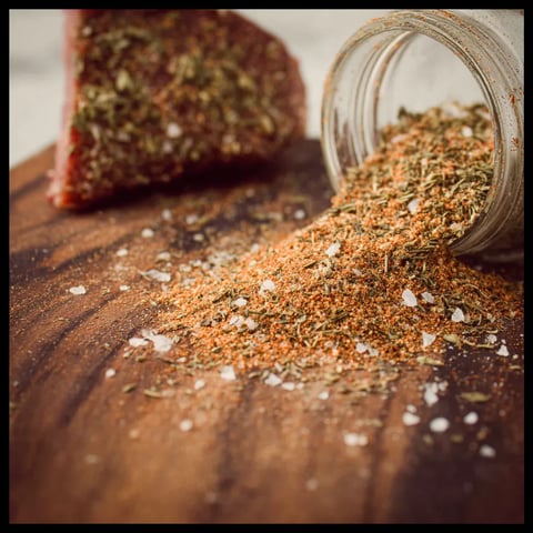 A jar of seasoning sits next to a piece of steak.